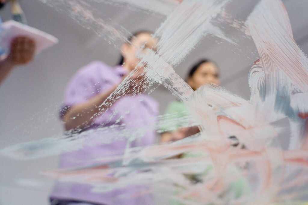 Women creating abstract art on glass with paint strokes, captured with selective focus.